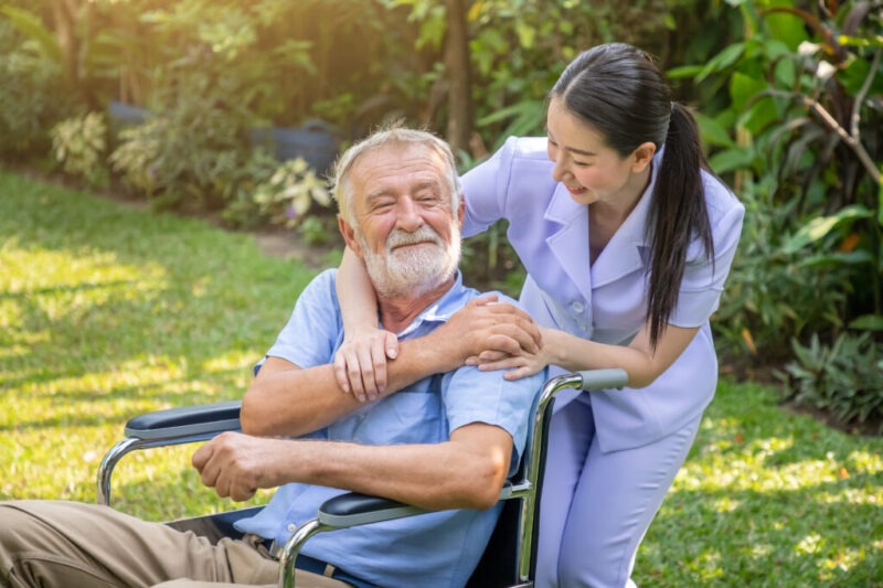 Caregiver supporting elderly man in wheelchair at nursing home garden